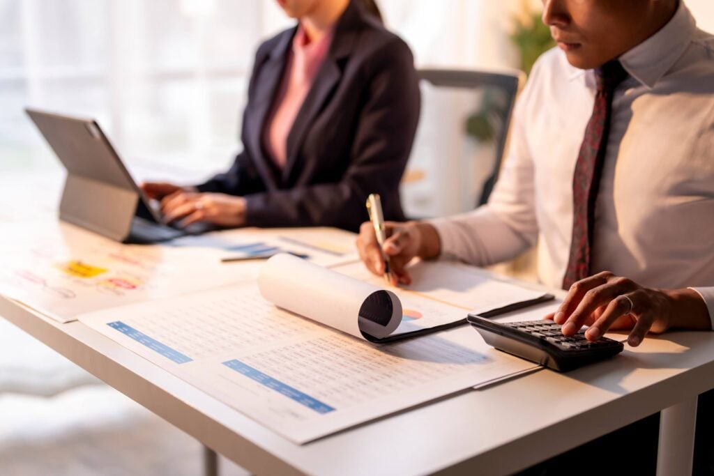 Two business professionals sit at a desk with financial documents, a calculator, and a laptop, working together in a bright office setting as they analyze data integration using the Sage Intacct API.
