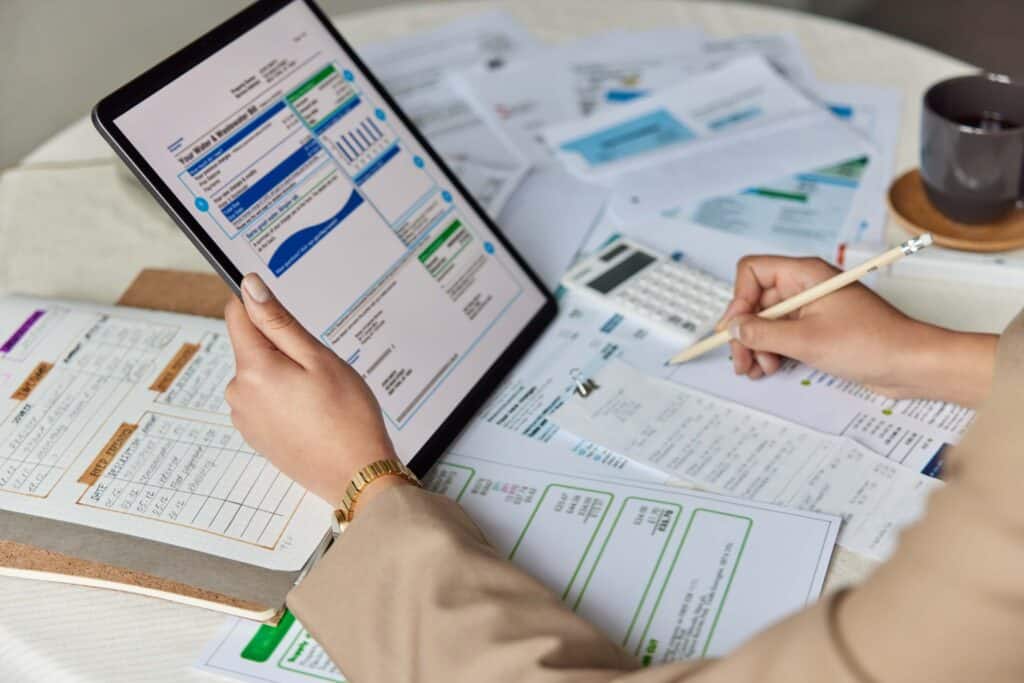 Person holding a tablet displaying graphs and data, surrounded by printed charts, documents, a clipboard, a pencil, and a calculator on a table—suggesting a work or finance-related task that could be streamlined using Beanworks by Quadient.