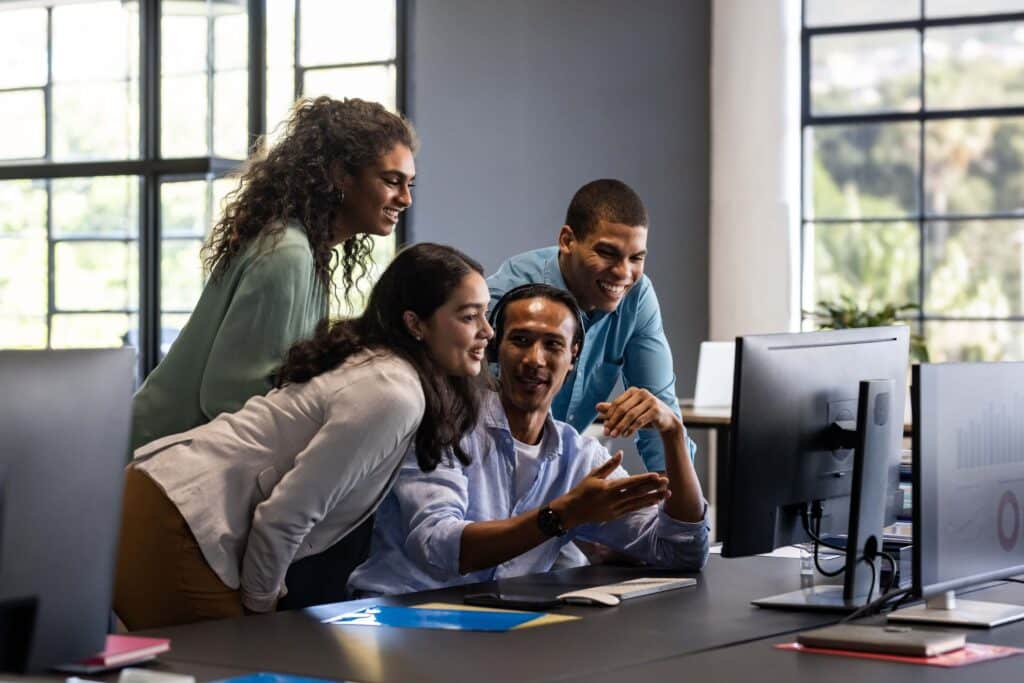 Four colleagues gather around a desk, smiling and looking at a computer monitor in a modern, bright office space as they collaborate on integrating the Sage Intacct API into their project.