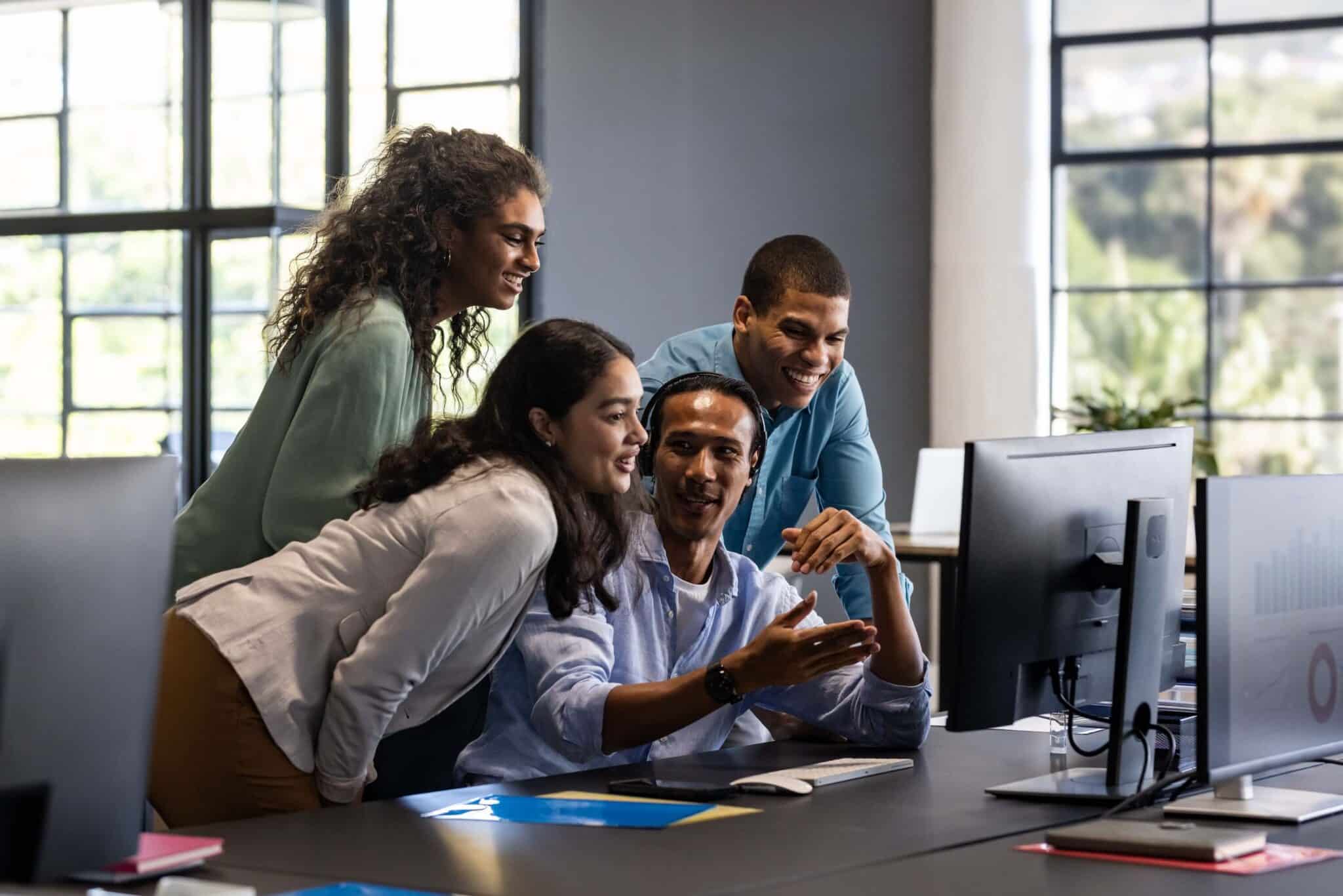 Four colleagues gather around a desk, smiling and looking at a computer monitor in a modern, bright office space as they collaborate on integrating the Sage Intacct API into their project.