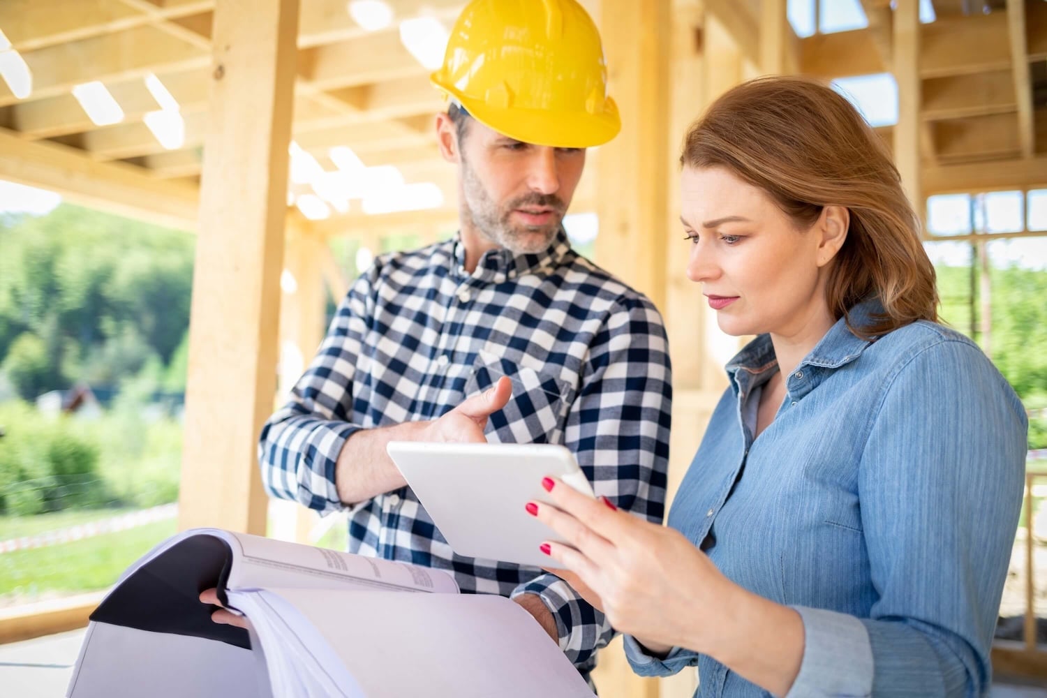 A man in a yellow hard hat and a woman with a tablet are discussing building plans at a construction site with wooden framing in the background, efficiently accessing project details through the Deltek Vantagepoint mobile app.