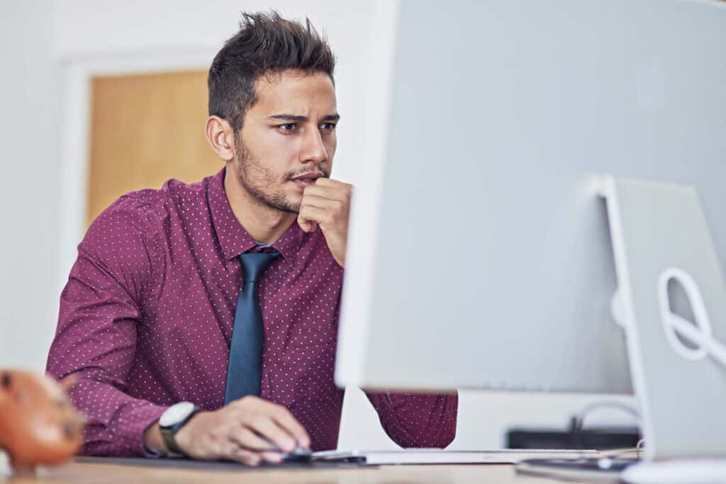 A man in a maroon shirt and tie sits at a desk, looking intently at a computer monitor with his hand on his chin, appearing focused and thoughtful as he reviews Deltek Vantagepoint custom reports.
