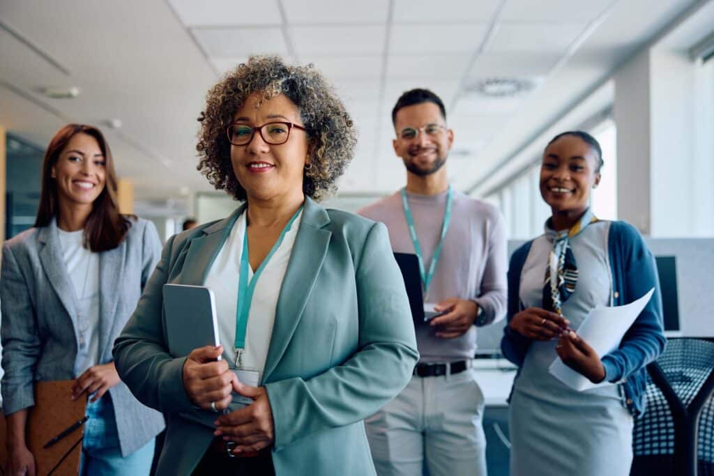 Four professionally dressed people stand in an office, smiling at the camera. The person in front holds a tablet—perhaps reviewing data through the Sage Intacct API—while others hold documents or a clipboard. They appear confident and collaborative.