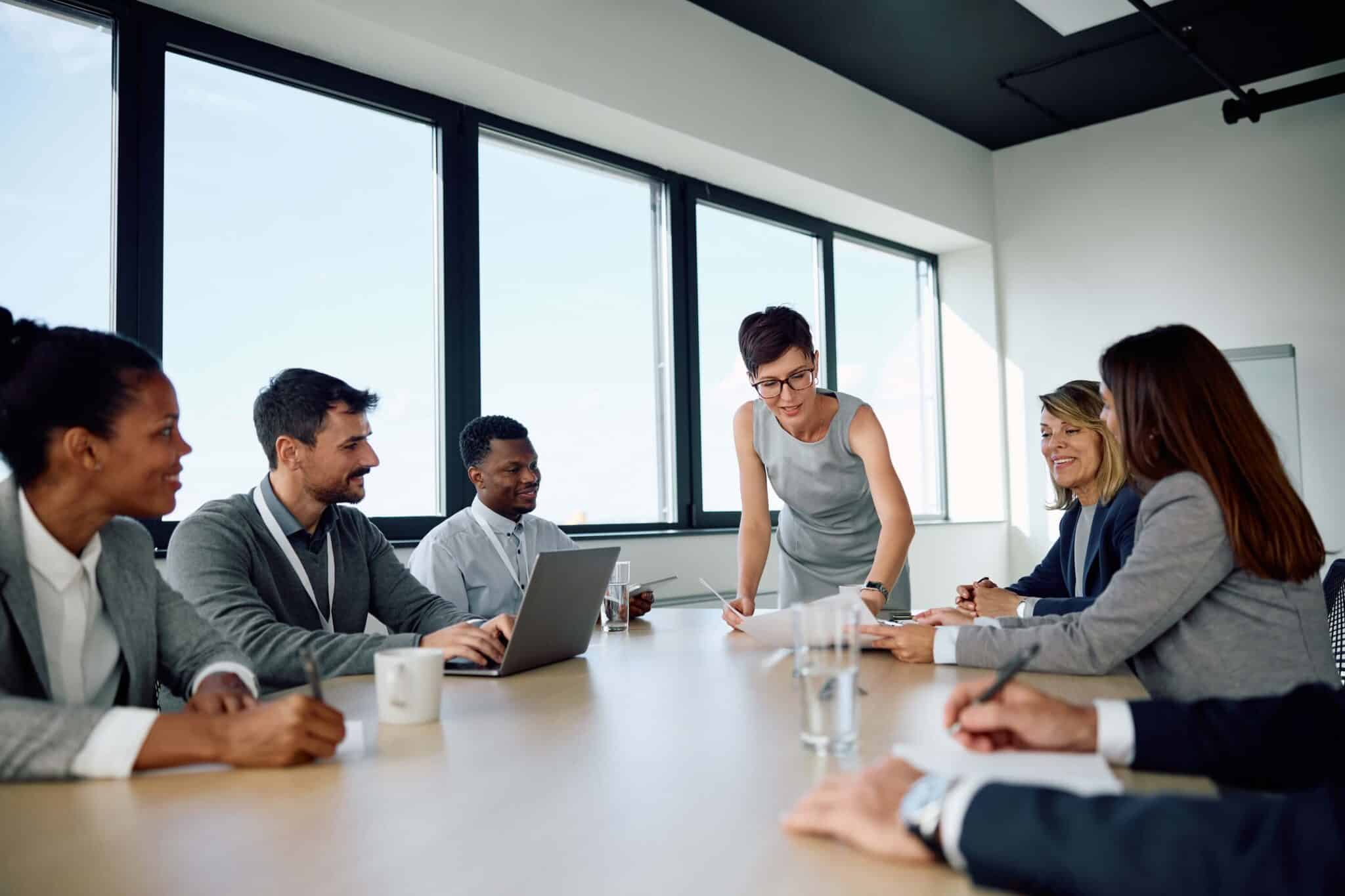 A group of professionals in business attire sit around a conference table, with one person standing and presenting deltek vantagepoint reports, in a modern office with large windows.
