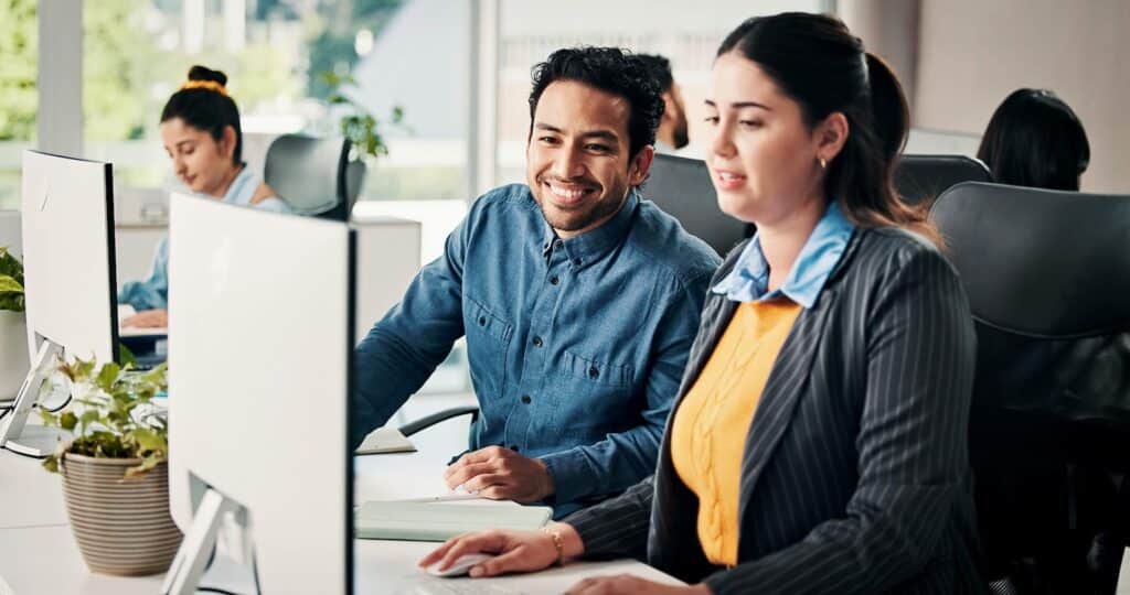 Two colleagues work together at computers in a modern office. One, smiling, looks at the other, who is focused on her screen exploring the Sage Intacct marketplace. Other employees work in the background. Plants and natural light add to the workspace.