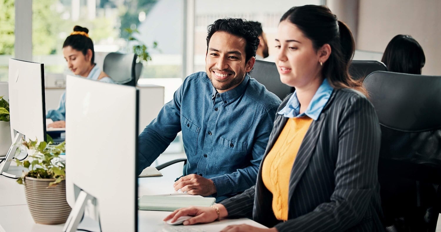 Two colleagues work together at computers in a modern office. One, smiling, looks at the other, who is focused on her screen exploring the Sage Intacct marketplace. Other employees work in the background. Plants and natural light add to the workspace.