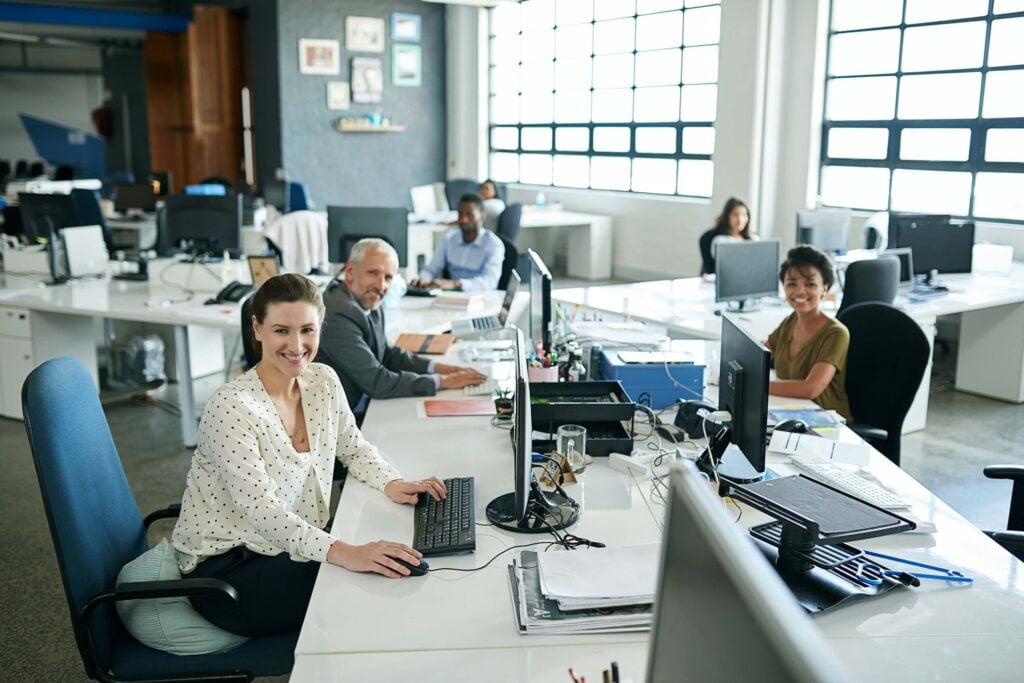 Five people sit at desks with computers in a bright, modern office. They are smiling and appear to be working together in an open-plan workspace with large windows and office equipment visible—perfectly capturing the collaborative spirit of businesses utilizing Quadient AP by Beanworks for streamlined accounts payable processes.