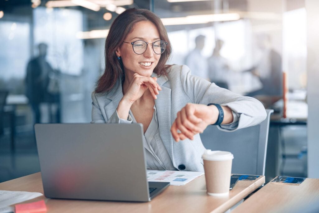 A woman in business attire sits at a desk with a laptop and coffee cup, smiling while looking at her smartwatch in a modern office setting, perhaps checking updates on replicon pricing or managing tasks related to replicon timesheet pricing.