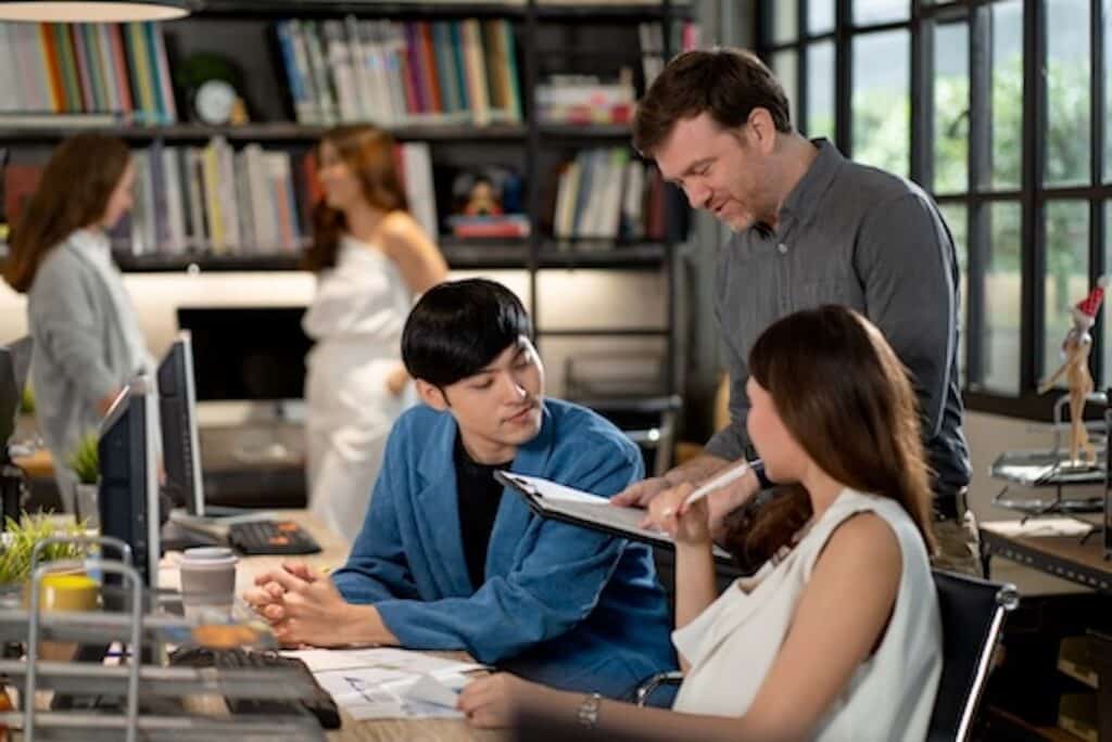 Three people sit and stand around a desk discussing documents in a modern office, demonstrating teamwork that reflects the collaborative spirit of Beanworks, while two others talk in the background near bookshelves and large windows.