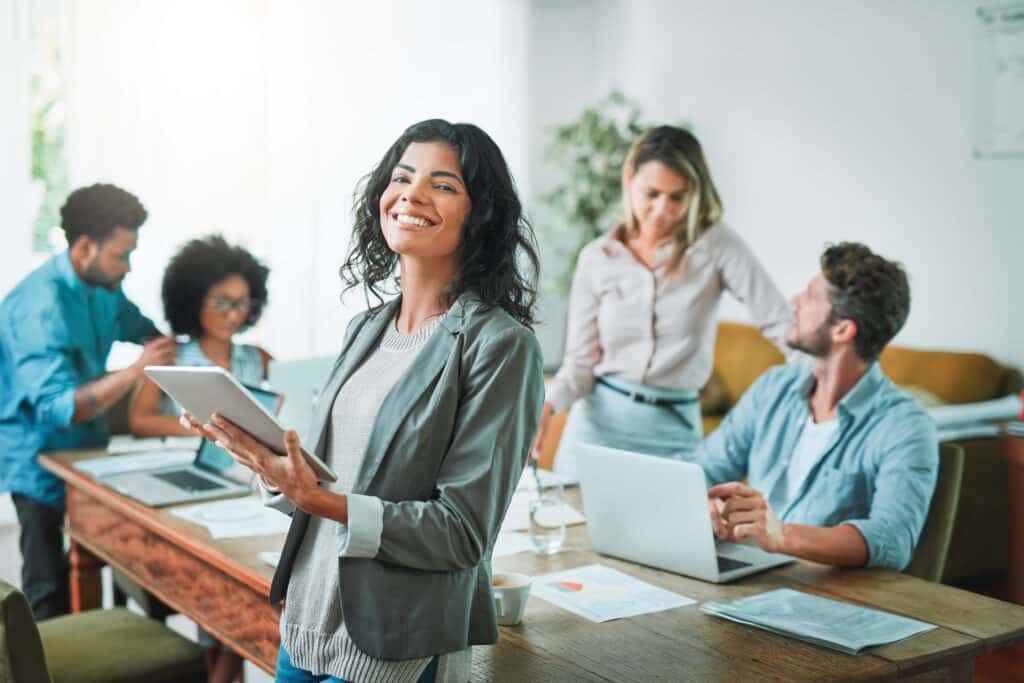 Smiling woman holding a tablet stands in front of a table where four colleagues collaborate with laptops and papers in a bright office setting, effortlessly reviewing Deltek Vantagepoint reports to streamline their workflow.