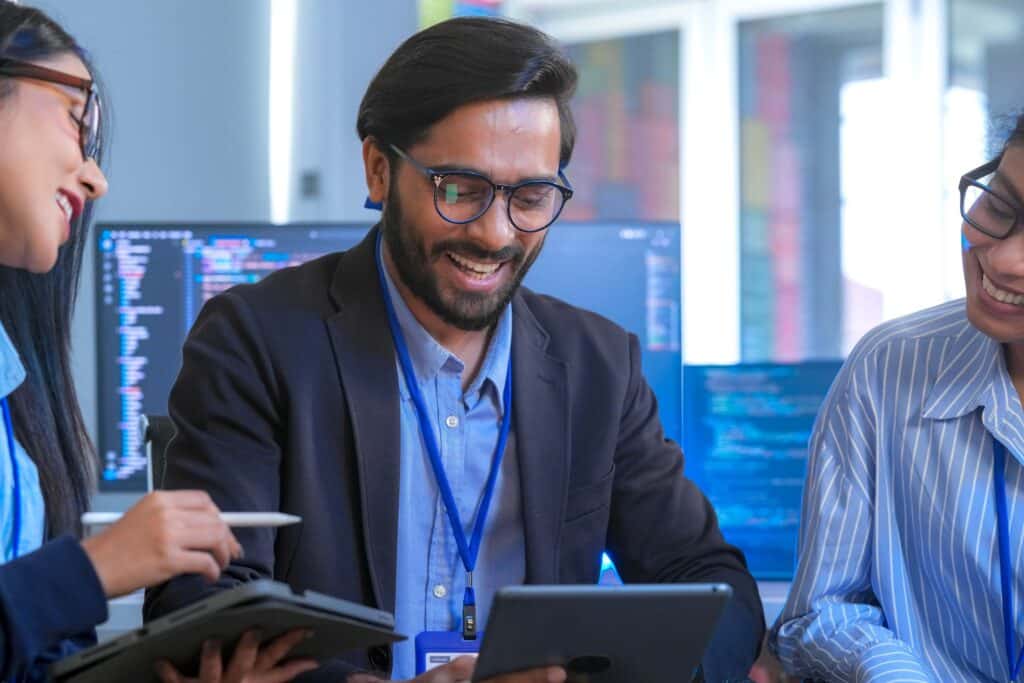 Three people wearing business attire smile and collaborate while looking at a tablet. Computer monitors with code are in the background, suggesting a tech or office environment where they may be discussing solutions such as the Sage Intacct API for seamless system integration.