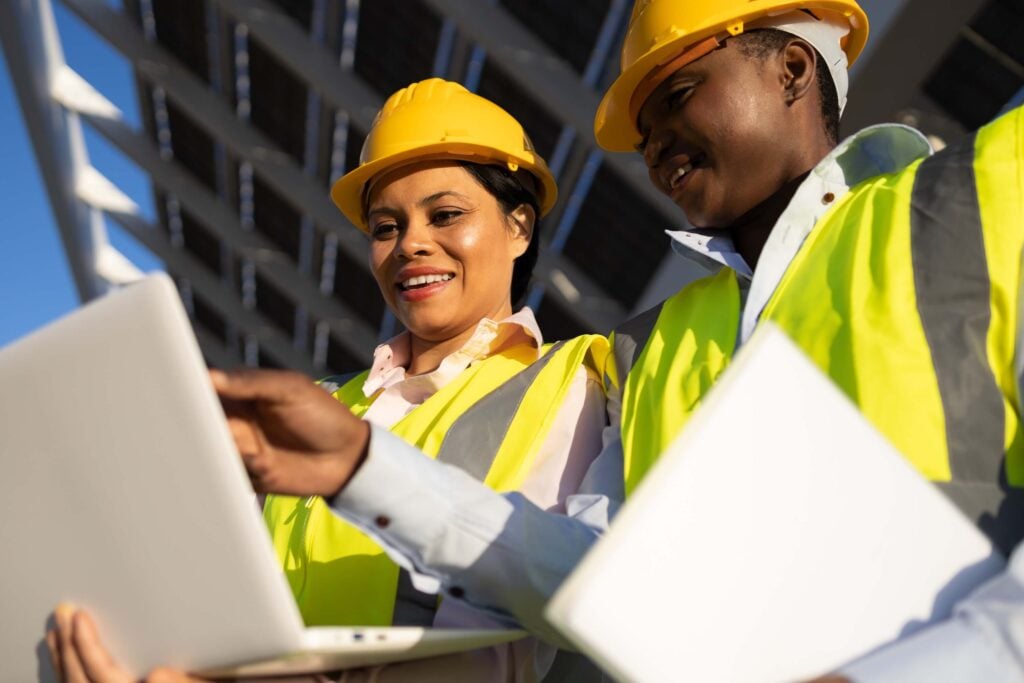 Two construction workers wearing yellow safety vests and hard hats smile while looking at a laptop outdoors, possibly at a construction or solar panel site, as they explore solutions from the Sage Intacct Marketplace to streamline their project management.
