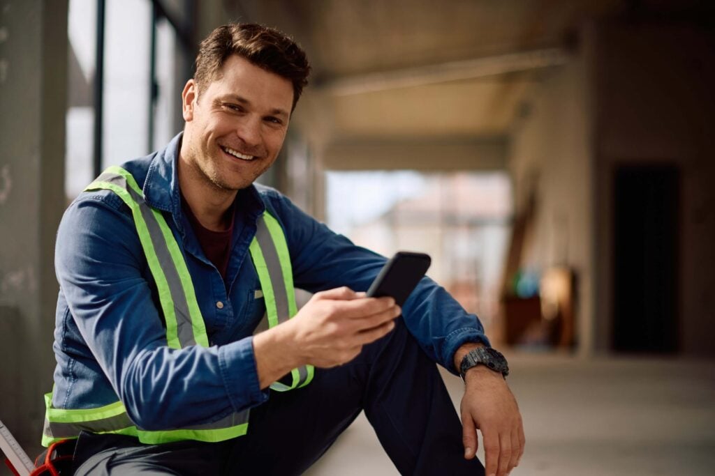 Smiling construction worker wearing a reflective safety vest sits indoors on site, holding a smartphone and looking at the camera—ready to manage tasks efficiently with the Deltek Vantagepoint mobile app.