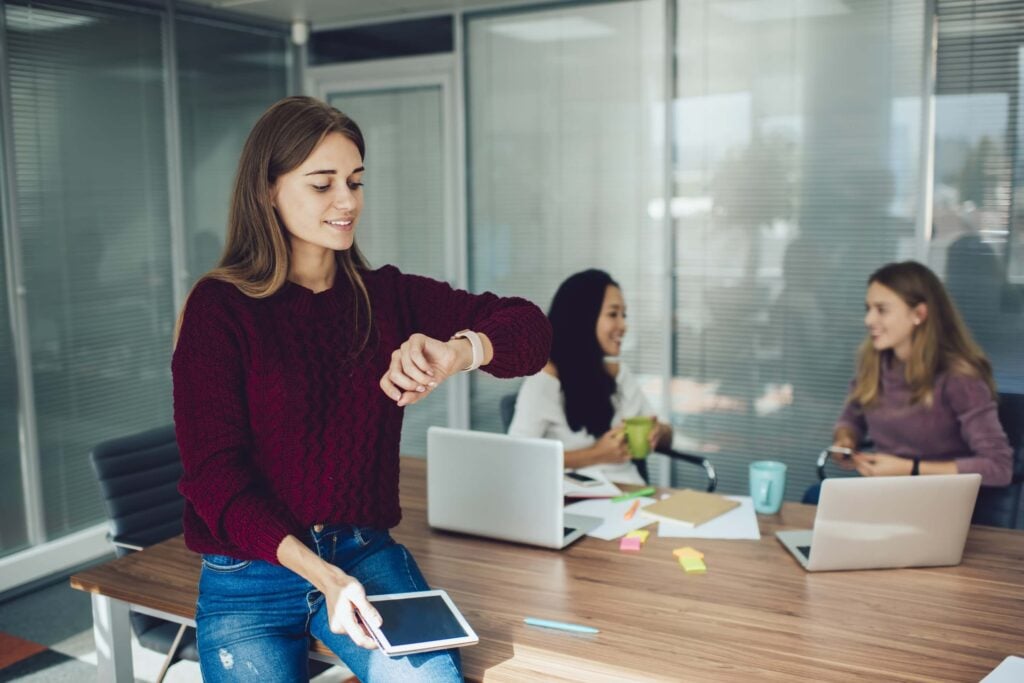 A woman in a maroon sweater checks her smartwatch while holding a tablet, perhaps reviewing replicon timesheet pricing for the team’s latest project. Two other women sit at a table with laptops, chatting in a modern office with glass walls.