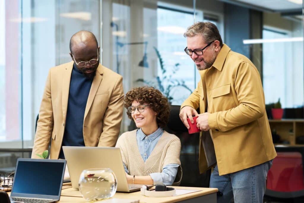 Three people in an office, two standing and one sitting at a desk with a laptop, are smiling and appear to be collaborating—perhaps discussing replicon pricing. There is a glass bowl on the desk and large windows in the background, creating an open and inviting atmosphere for teamwork.