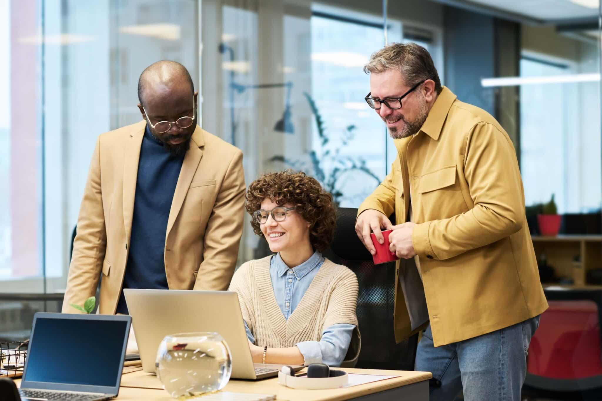 Three people in an office, two standing and one sitting at a desk with a laptop, are smiling and appear to be collaborating—perhaps discussing replicon pricing. There is a glass bowl on the desk and large windows in the background, creating an open and inviting atmosphere for teamwork.