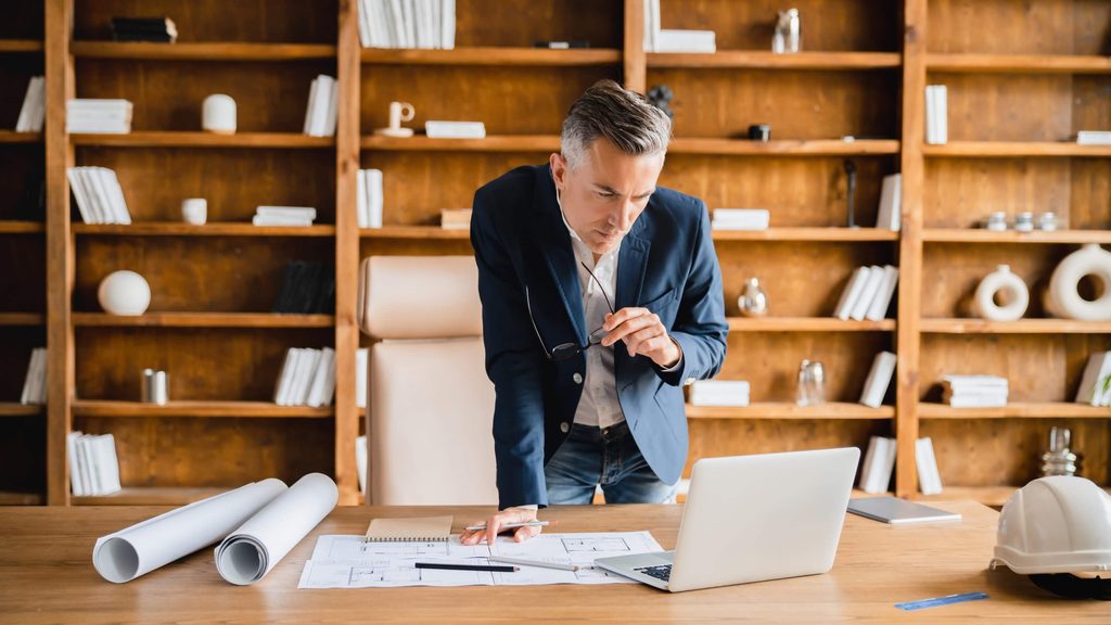 A man in a blazer stands at a desk with architectural plans, looking at a laptop running Deltek Vantagepoint 2026.2 in a modern office with shelves and books in the background, possibly using Ask Dela to streamline his workflow.