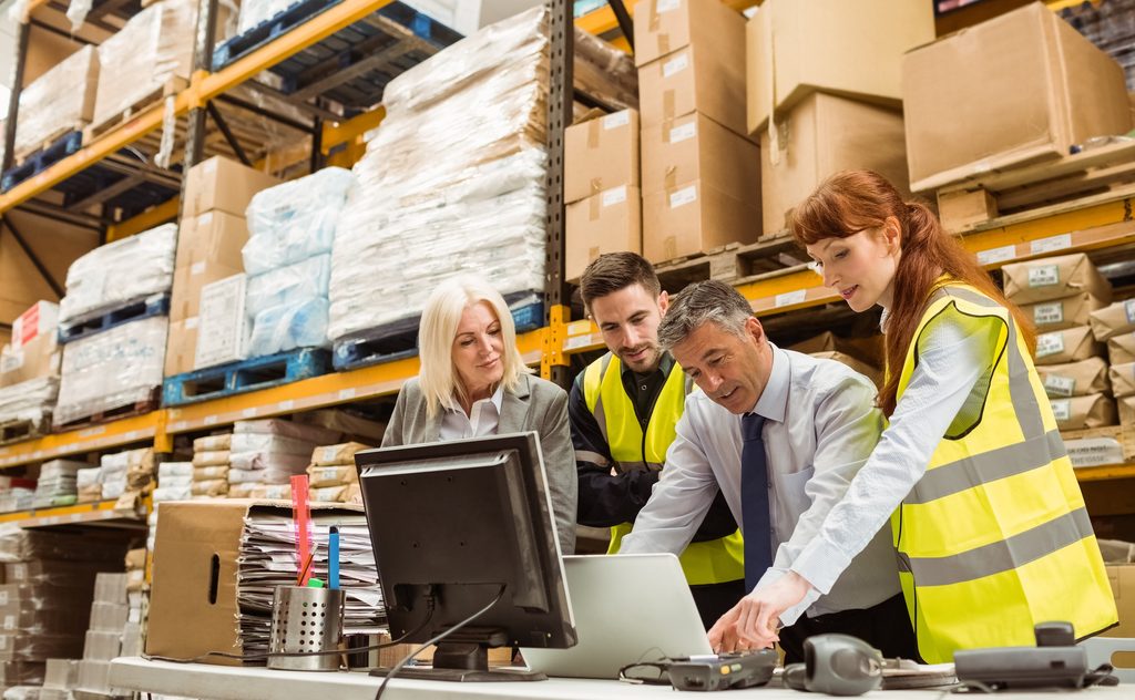 Four people in a warehouse, two wearing yellow safety vests, gather around a computer and paperwork, discussing something—perhaps reviewing inventory with sage 100 warehouse management. Shelves with boxes and pallets are stacked in the background.