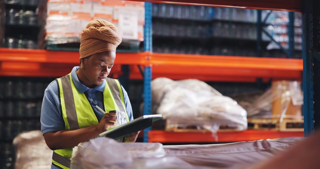 A person in a yellow safety vest writes on a clipboard in a warehouse filled with shelves and large wrapped packages, efficiently overseeing inventory with sage 100 warehouse management solutions.