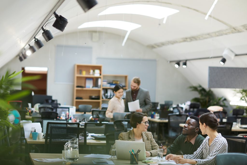 Open office space with people collaborating in small groups at desks, surrounded by chairs, plants, and shelves under a curved white ceiling—an inspiring environment ideal for teamwork and projects such as a Sage 100 upgrade.