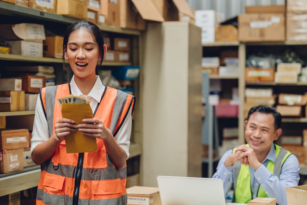 A woman in a safety vest smiles while holding cash and an envelope in a warehouse, demonstrating the efficiency that sage 100 warehouse management can bring to daily operations. In the background, a man in a safety vest sits at a desk with a laptop, smiling as he works. Shelves with boxes line the walls.