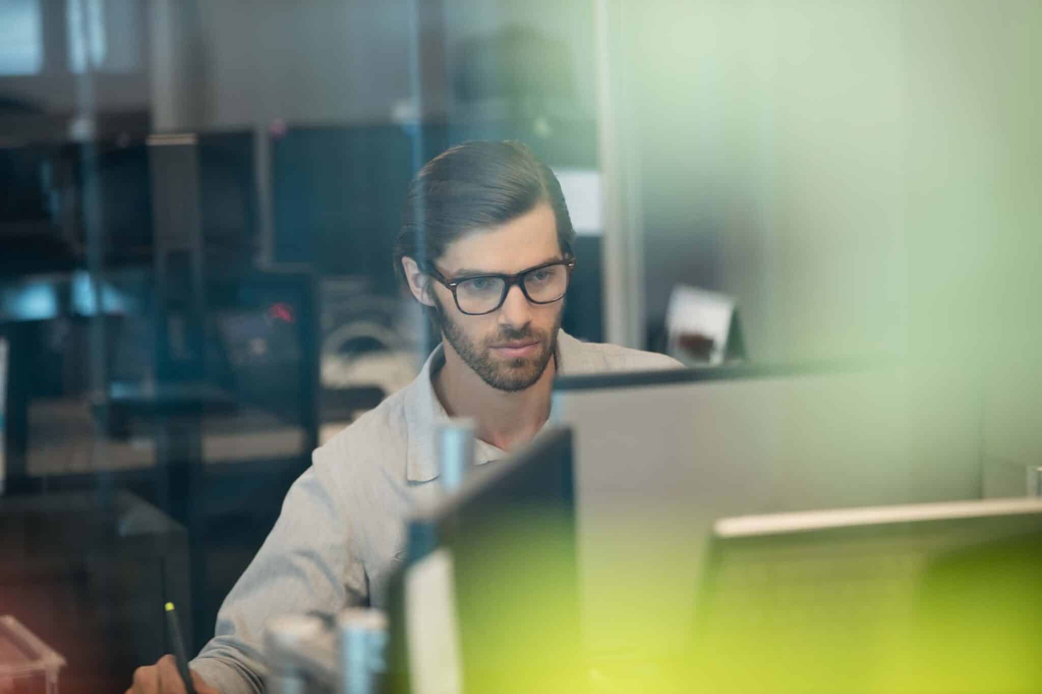 A man with glasses and a beard works at a computer in a modern office, focused intently on the screen as he develops sage 100 integrations. The foreground is blurred with green hues.