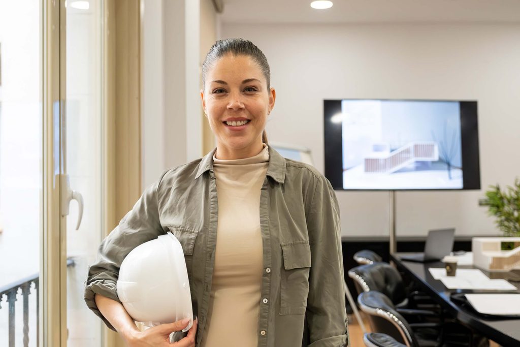 Woman smiling in an office, holding a white hard hat. A presentation with an architectural design and insights from Deltek Vantagepoint 2026.2 is visible on a screen in the background, while Ask Dela support enhances her workflow.