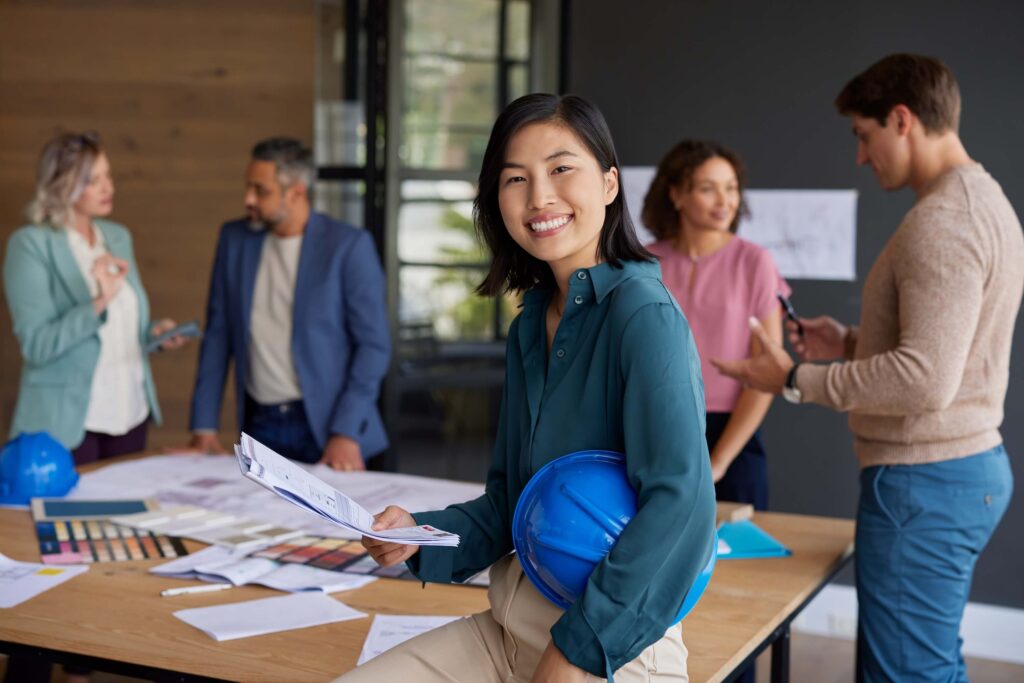 A woman holding blueprints and a blue hard hat smiles at the camera, while four colleagues discuss work and architectural plans around a table in a modern office—efficiently collaborating with Deltek Vantagepoint 2026.2 and leveraging Ask Dela for real-time insights.