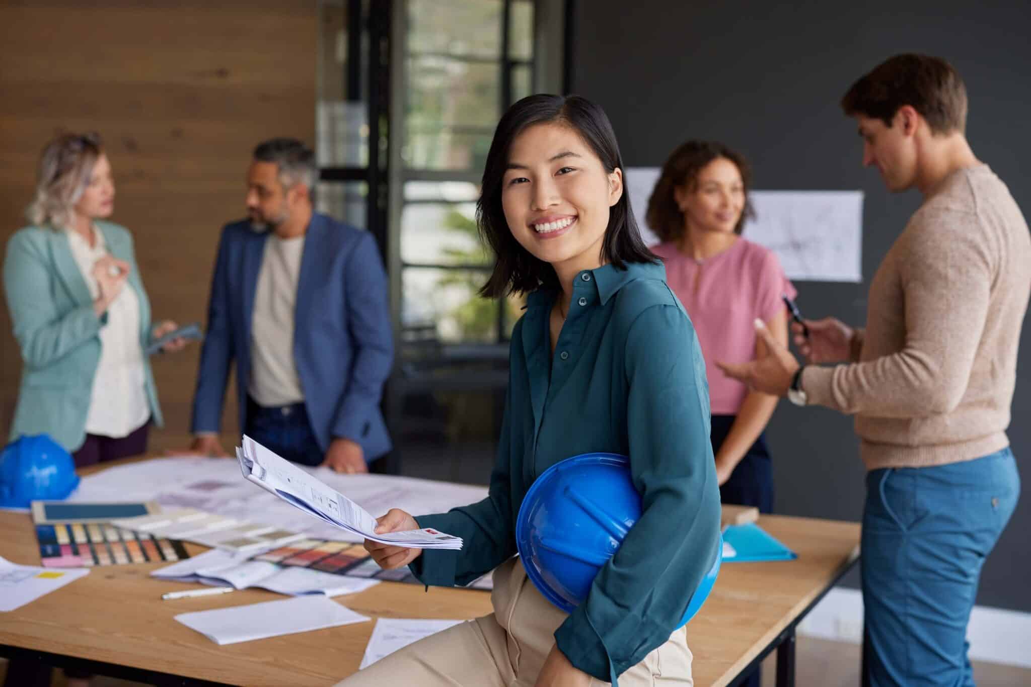 A woman holding blueprints and a blue hard hat smiles at the camera, while four colleagues discuss work and architectural plans around a table in a modern office—efficiently collaborating with Deltek Vantagepoint 2026.2 and leveraging Ask Dela for real-time insights.