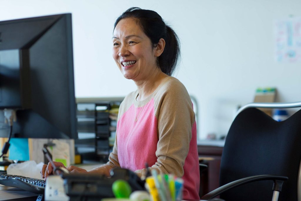 A woman in a pink and beige top smiles while working at her desk in an office, seamlessly managing tasks with the help of Sage 100 integration. There is a computer monitor, office supplies, and a black chair visible. The background is softly blurred.