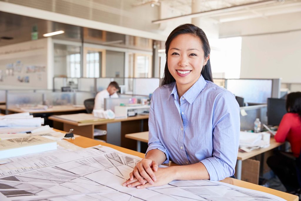Woman smiling and sitting at a desk covered with architectural blueprints in a modern office, efficiently managing projects using Deltek Vantagepoint 2026.2, while coworkers work collaboratively in the background. With Ask Dela at her fingertips, she navigates her tasks seamlessly and stays ahead in every aspect of the design process.
