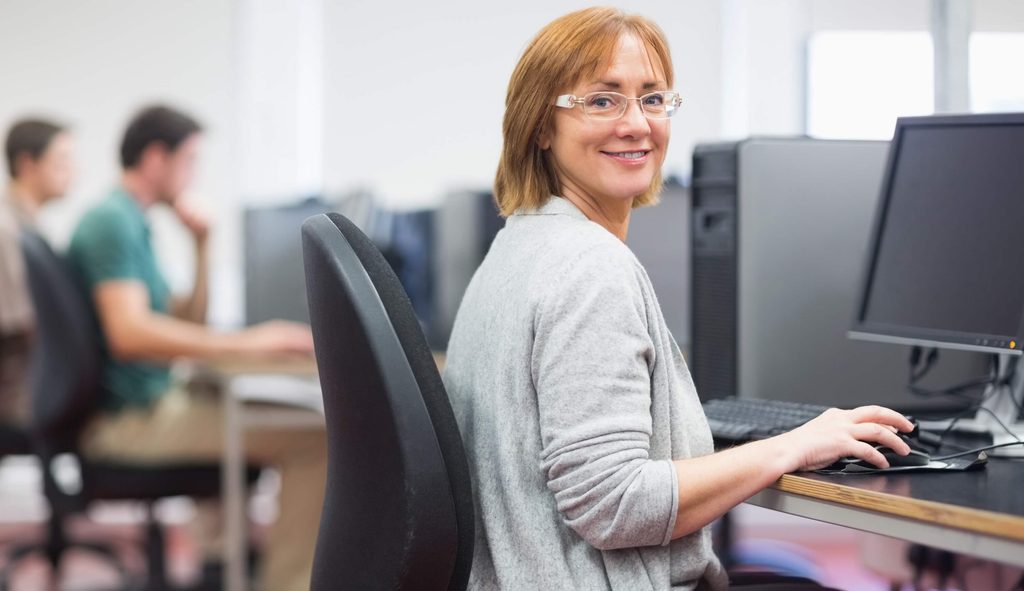 A woman wearing glasses smiles at the camera while using a desktop computer in an office, where she is focused on Sage 100 integration. Other people work at computers in the background.