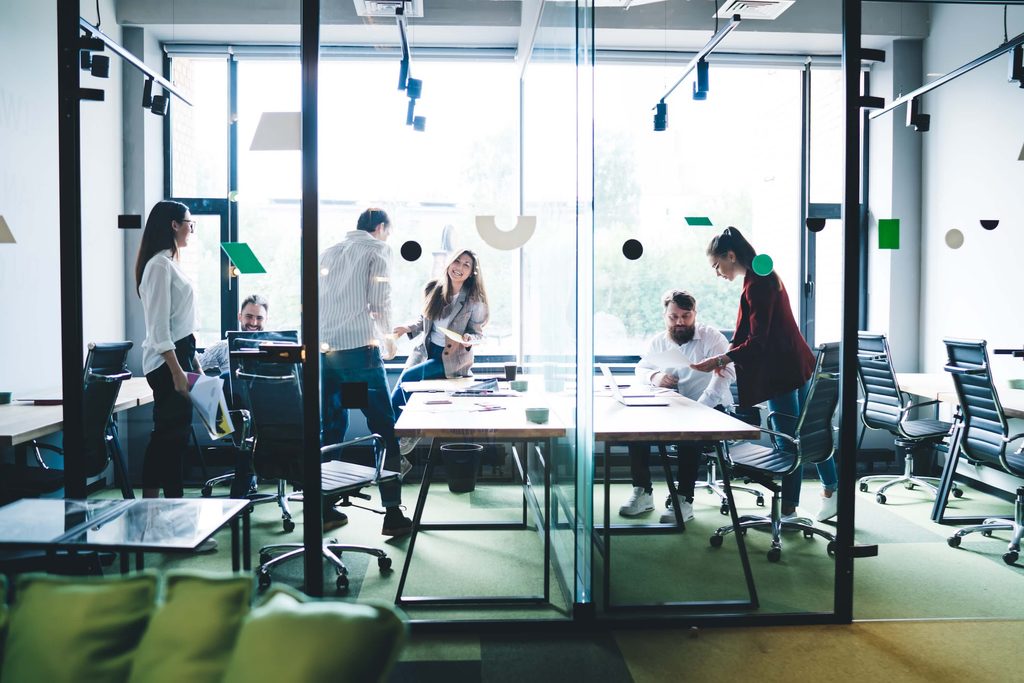 Six people collaborate in a modern glass-walled office, standing and sitting around tables with papers and laptops, discussing solutions such as Sage 100 integration under bright natural light from large windows.