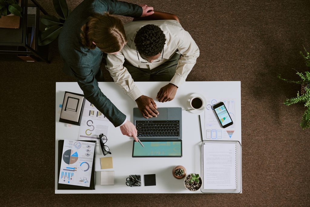 Two people sit at a desk with a laptop, documents, charts, smartphone, coffee, and plants, collaborating on a sage 100 upgrade and pointing at the laptop screen in an office setting.